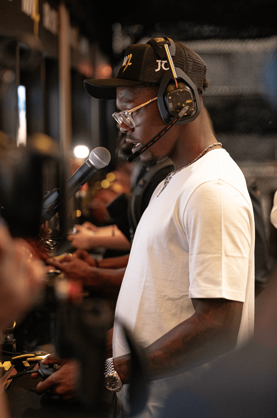 A person wearing a black cap with "JC" logo, headphones, and a white shirt speaking into a microphone on a stage.