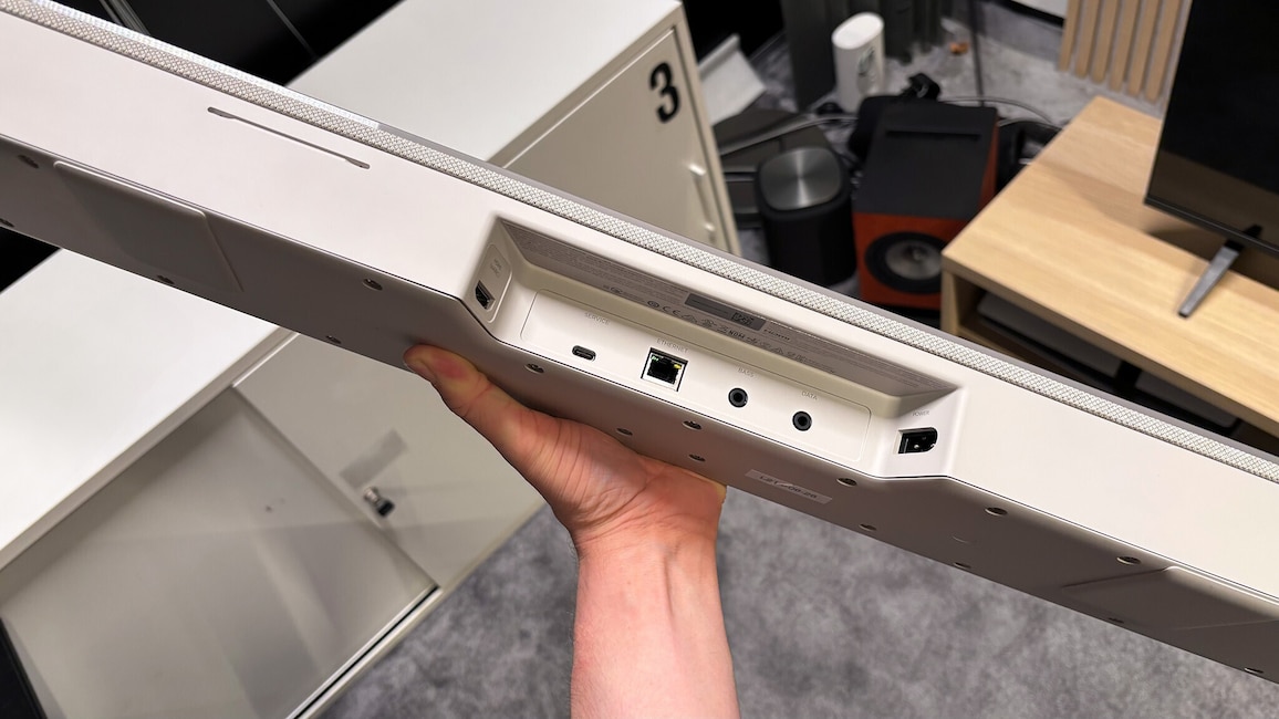 Eine Person hält die Bose Lifestyle Ultra Soundbar mit der Rückseite nach oben. Sichtbar sind die Anschlüsse für HDMI eARC, Ethernet, Service, Stromversorgung sowie die Verbindungen zum Subwoofer und weiteren Komponenten.