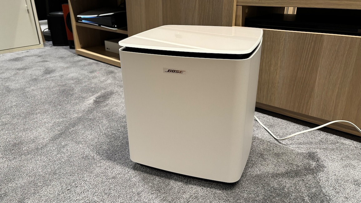 Nahaufnahme des kabellosen Bose Lifesyle Ultra Subwoofers. Das würfelförmige Gehäuse mit abgerundeten Kanten und glänzender Oberseite steht neben einem TV-Möbel.