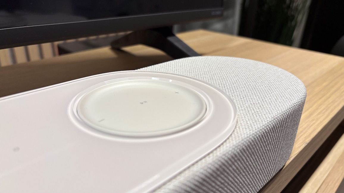 Nahaufnahme der Bose Lifestyle Ultra Soundbar. Zu sehen sind die helle Stoffbespannung, die glänzende Glasoberfläche und das große berührungsempfindliche Lautstärkerad auf der Oberseite.