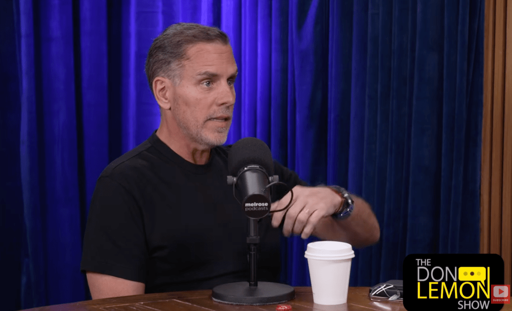 A man with gray hair speaking into a microphone on a stage with a blue curtain backdrop, wearing a black t-shirt with the Melrose Studios logo.