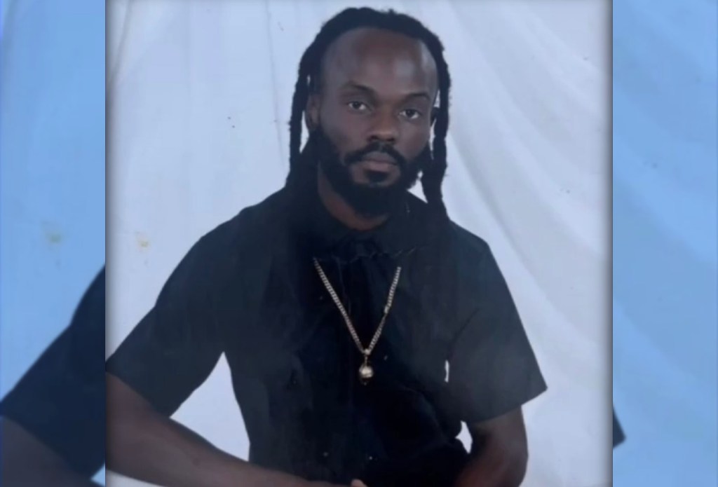 A man with dreadlocks wearing a black shirt and necklace, looking directly at the camera.