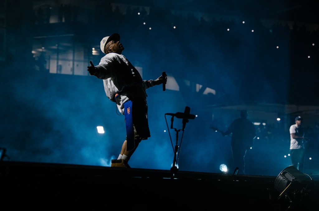 A person in a dark outfit and hat performing on a dimly lit stage, with spotlights and a microphone stand visible.