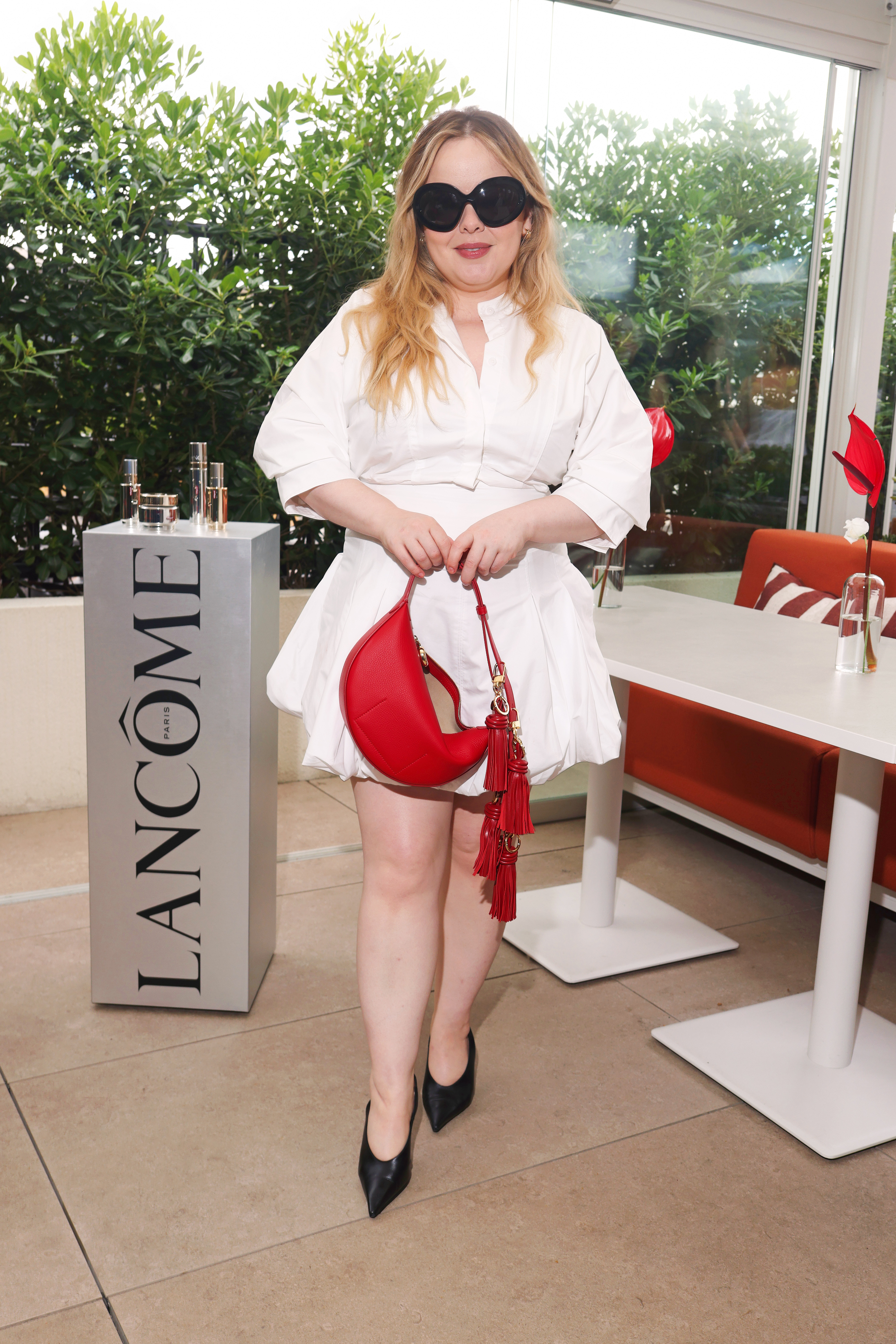 Nicola Coughlan wears a white bubble hem dress with black pointed-toe heels, a red bag and oversized sunglasses.