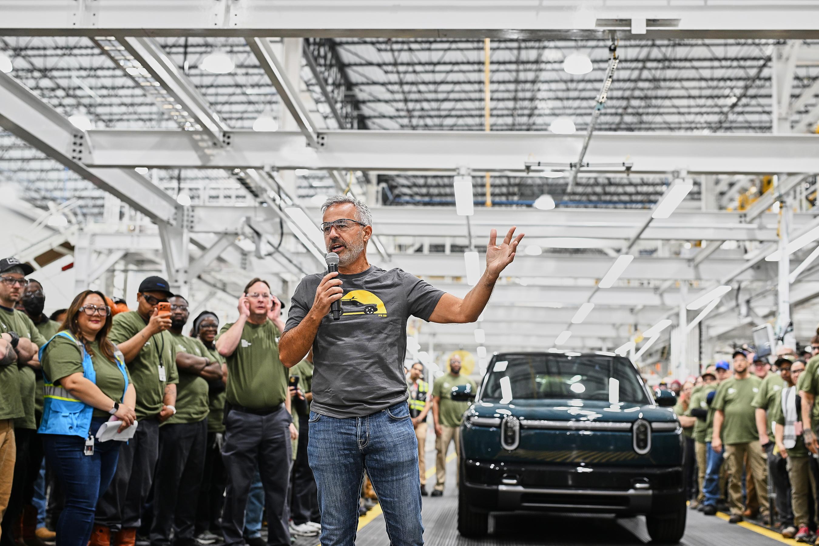 A man in a grey t-shirt and jeans holds a microphone and stands in front of a blue Rivian r2 inside of a huge, bright  factory space and speaks to a crowd of people who are mostly wearing green t-shirts.
