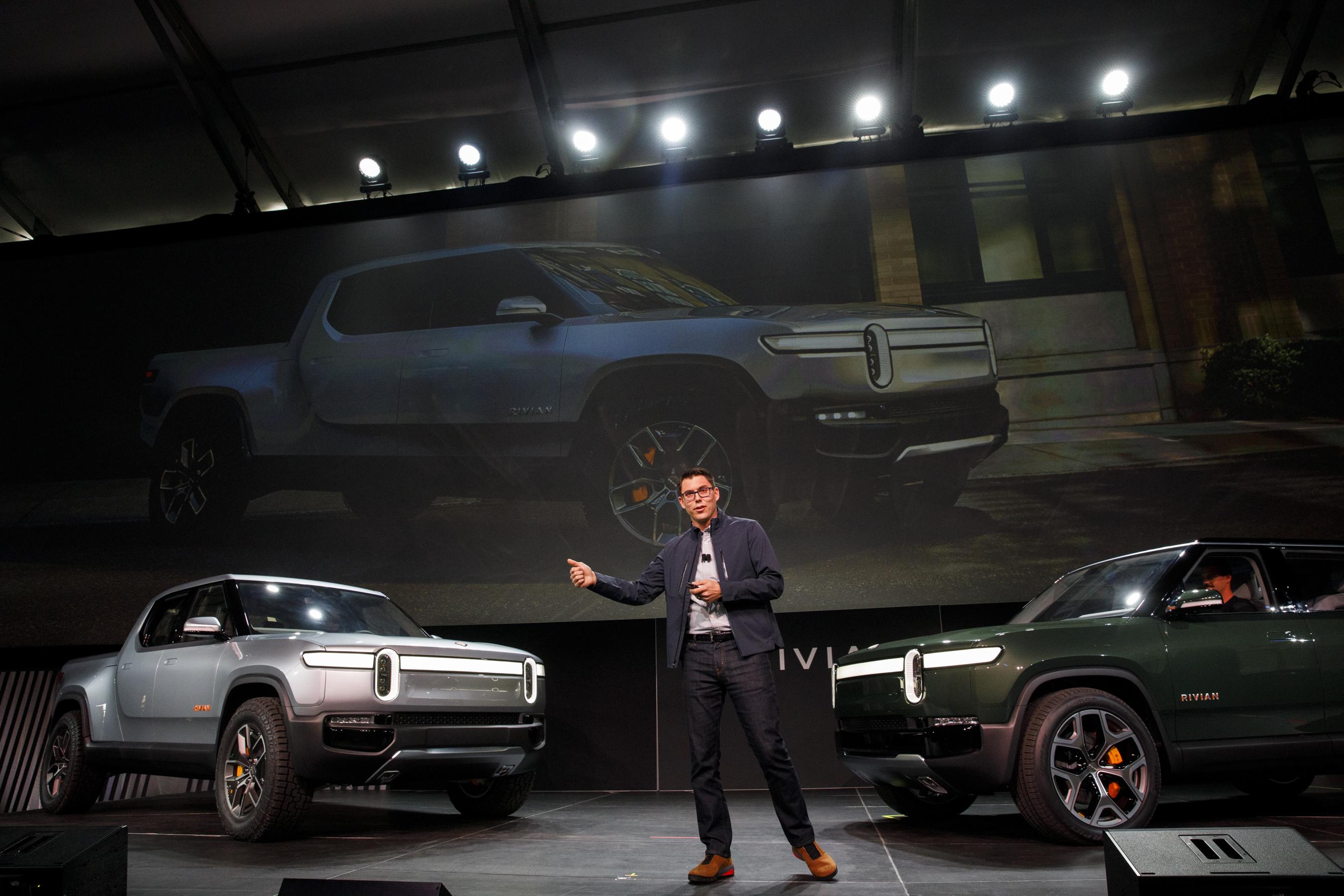 A man with glasses stands on stage with two Rivian vehicles facing him. 