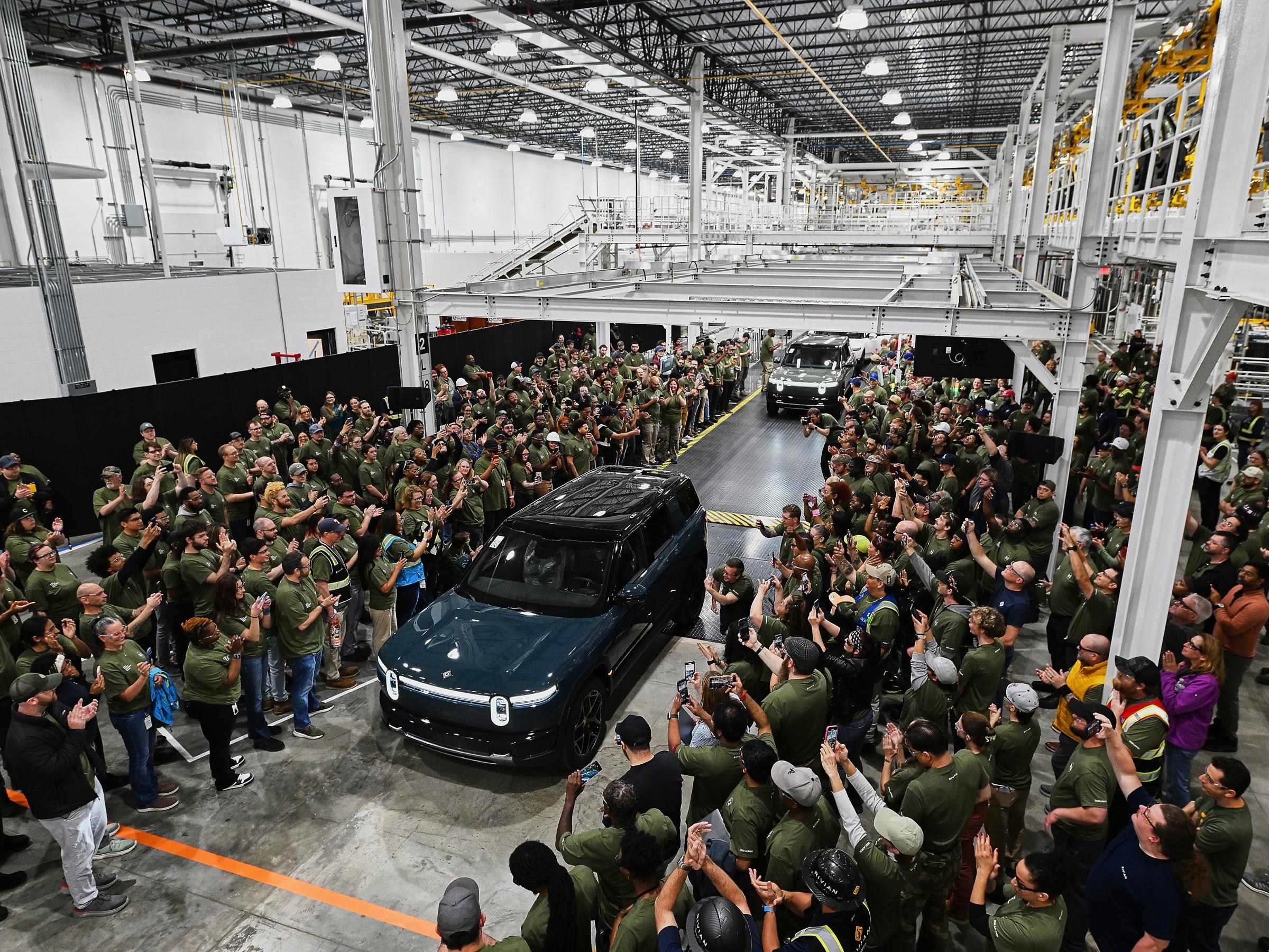 Wide view of a dark blue Rivian driving down a narrow path of the factory floor with people gathered on either side.&nbsp;