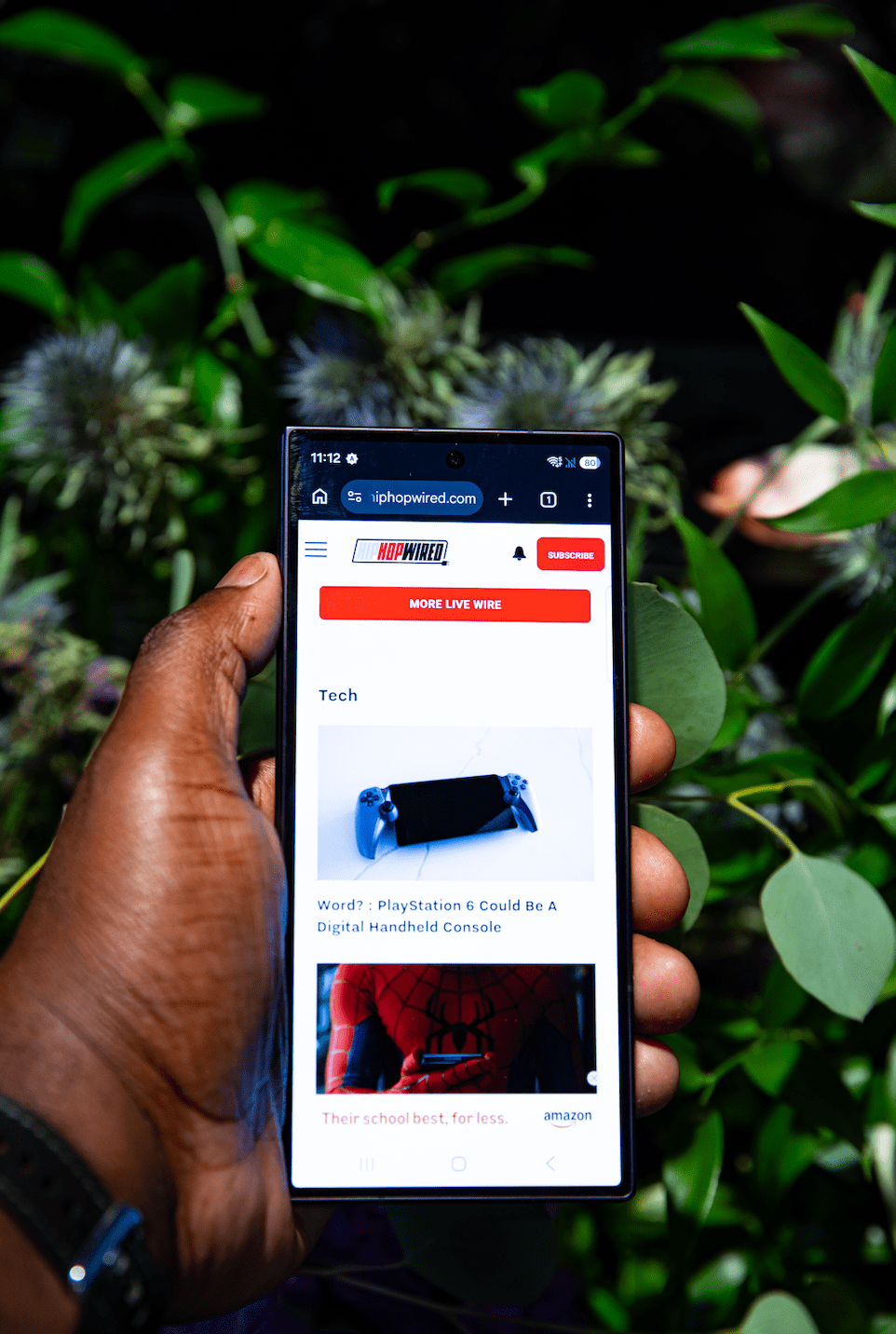 A hand holding a smartphone displaying a website with a PlayStation 6 controller image and text about it potentially being a digital handheld console.
