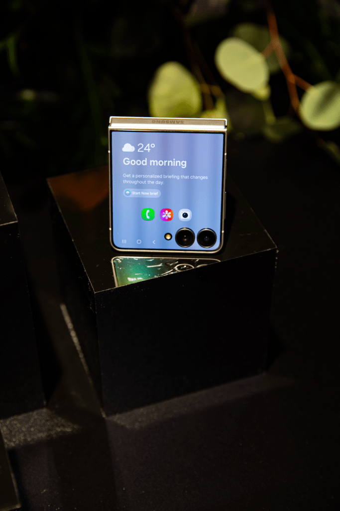 A smartphone displaying the time and weather information on a dark background with blurred greenery in the background.
