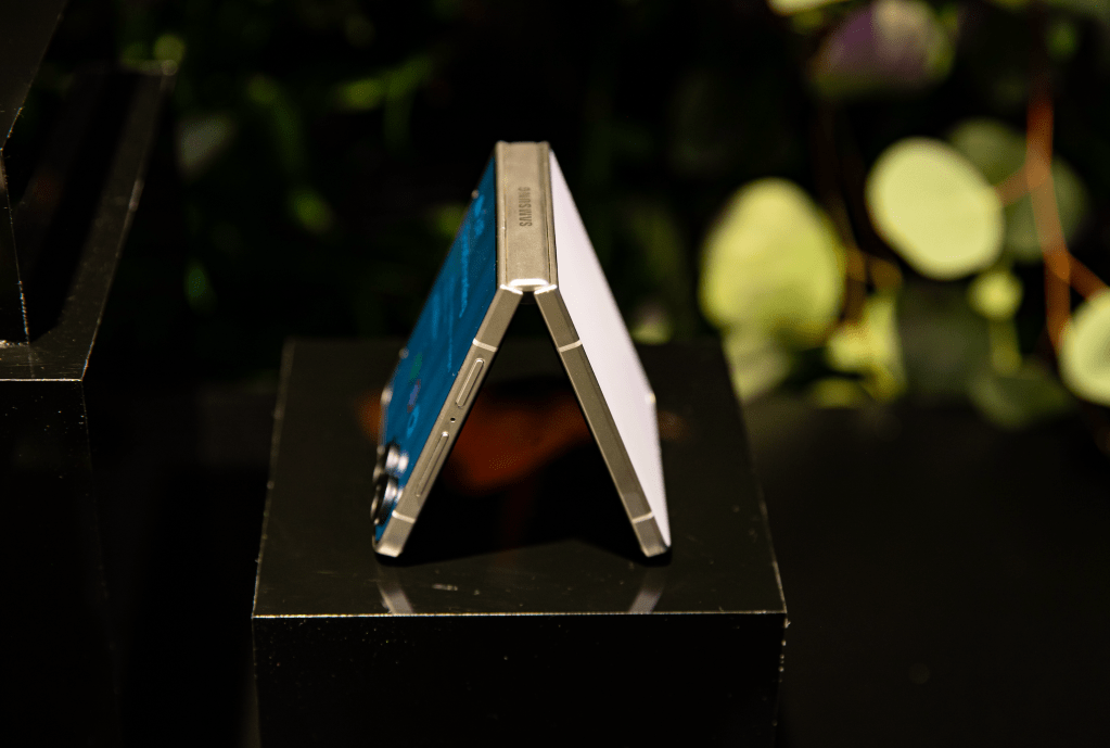 A silver-colored electronic device with a folding display, standing on a black surface against a blurred natural background.
