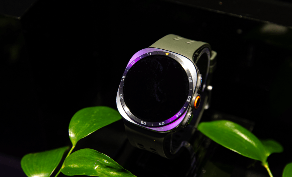 A black and green smartwatch with a purple and yellow dial face, resting on green leaves against a dark background.