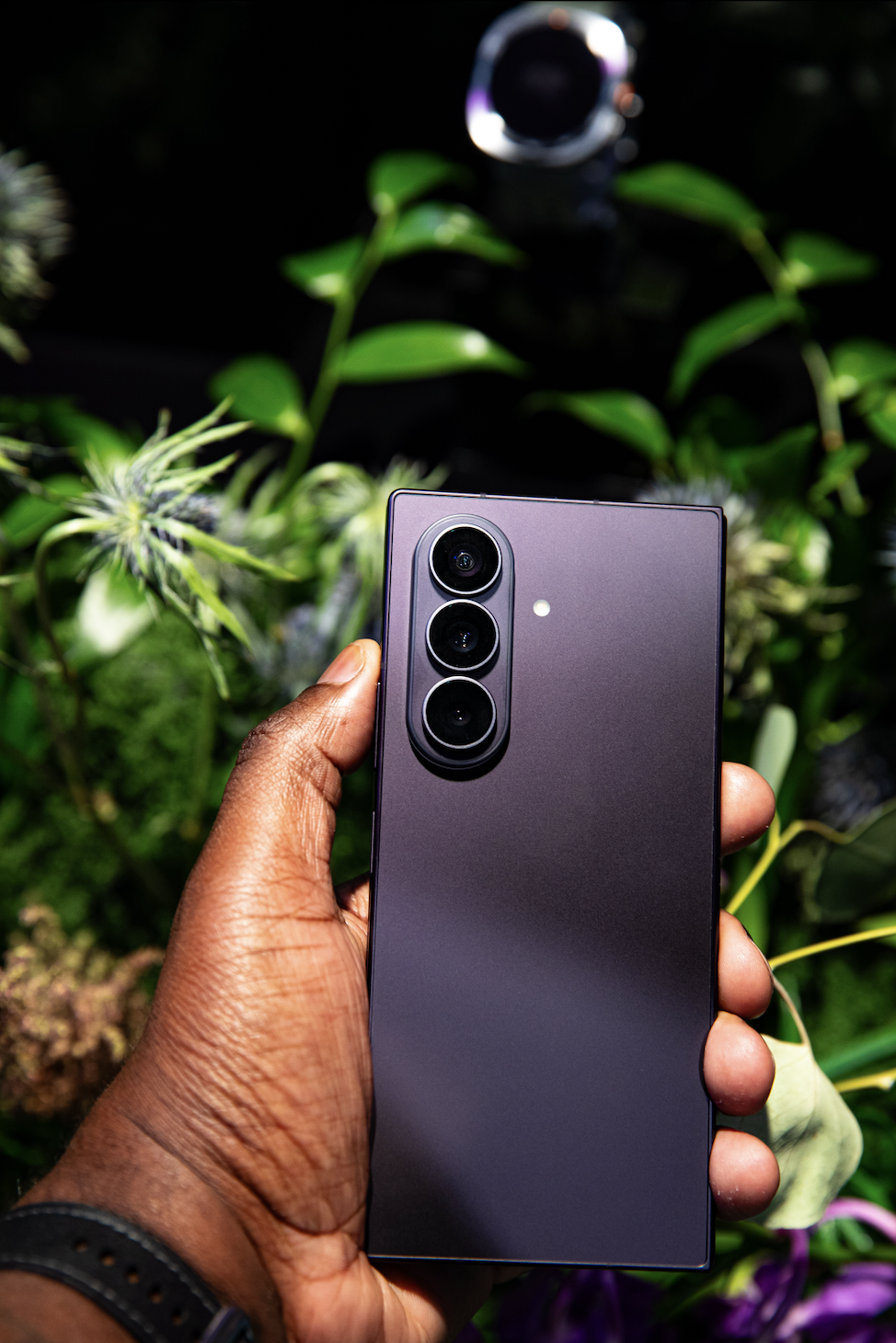 A hand holding a black smartphone with a triple camera lens against a backdrop of lush green foliage.