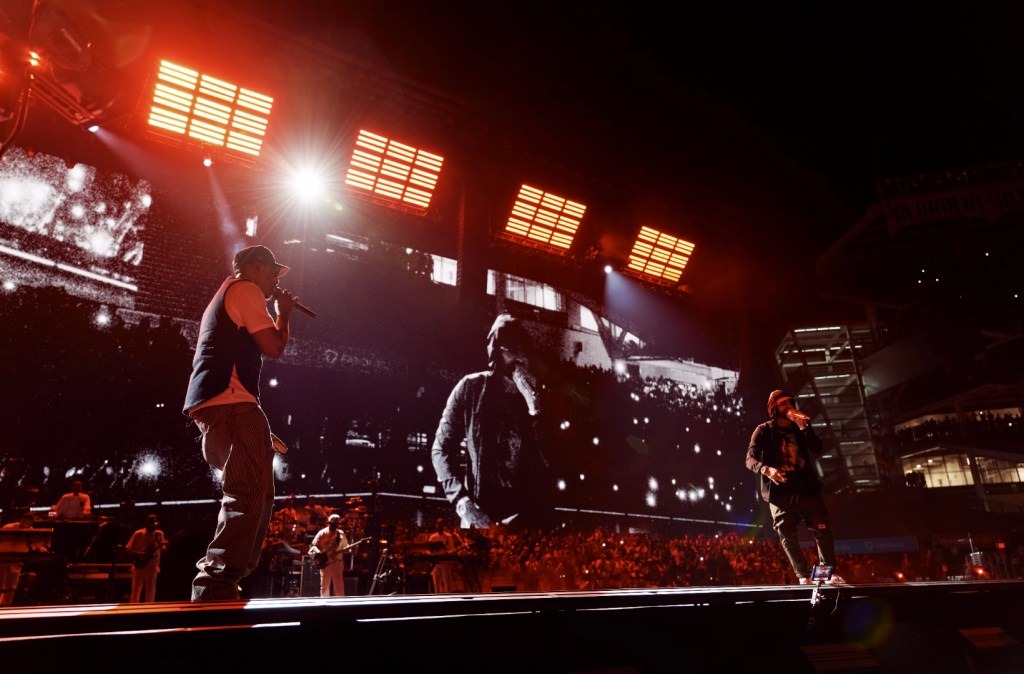 A concert stage at night, with performers on stage and a large crowd in the audience. The stage is illuminated by bright lights and spotlights, creating a dramatic atmosphere.