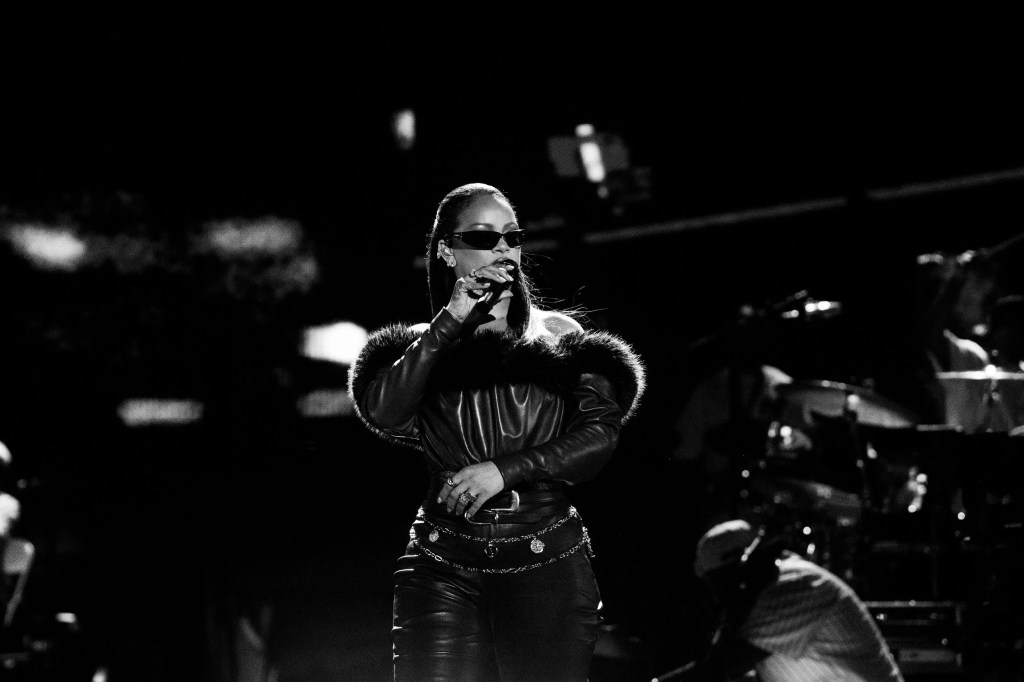 A person wearing sunglasses and a fur-trimmed jacket performing on a stage in a dark setting.