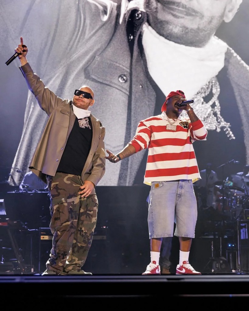 Two performers on stage, one wearing camouflage pants and a black top, the other in a red and white striped shirt, both holding microphones and performing.