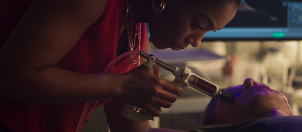 A person in a red shirt holds a gun to another person's head in a dimly lit room.