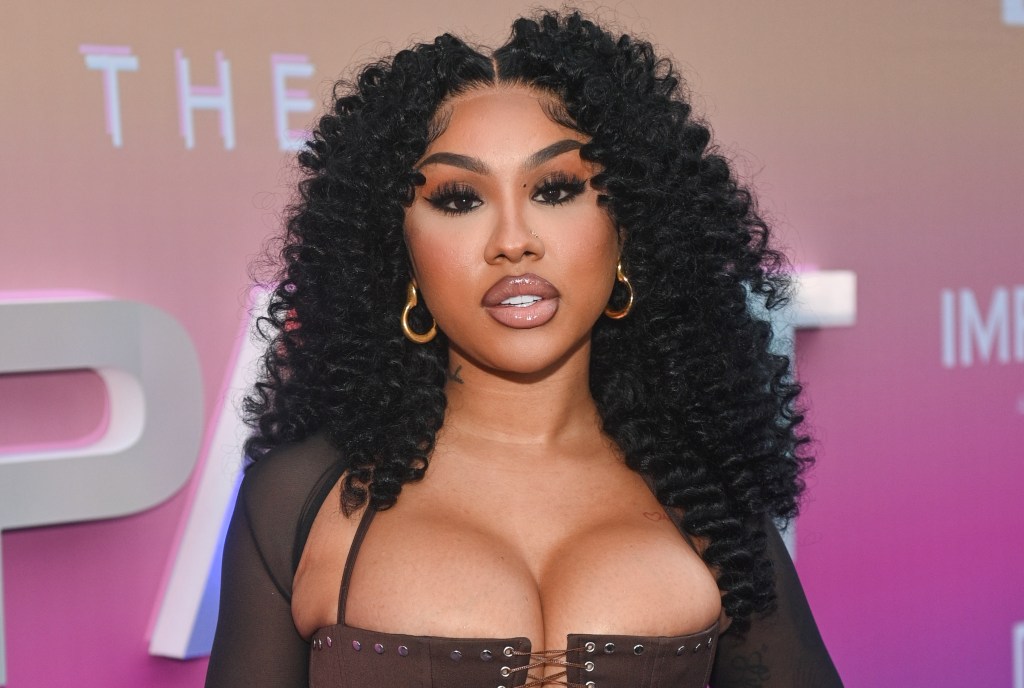A woman with curly black hair wearing a black outfit and large hoop earrings poses in front of a pink and white backdrop with the text "THE IMPACT ATLANTA".