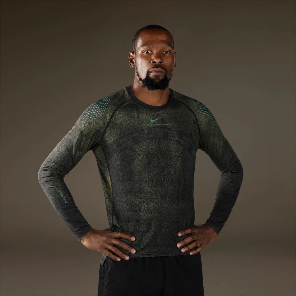 A man with a beard wearing a green Nike athletic shirt stands with his hands on his hips against a dark background.