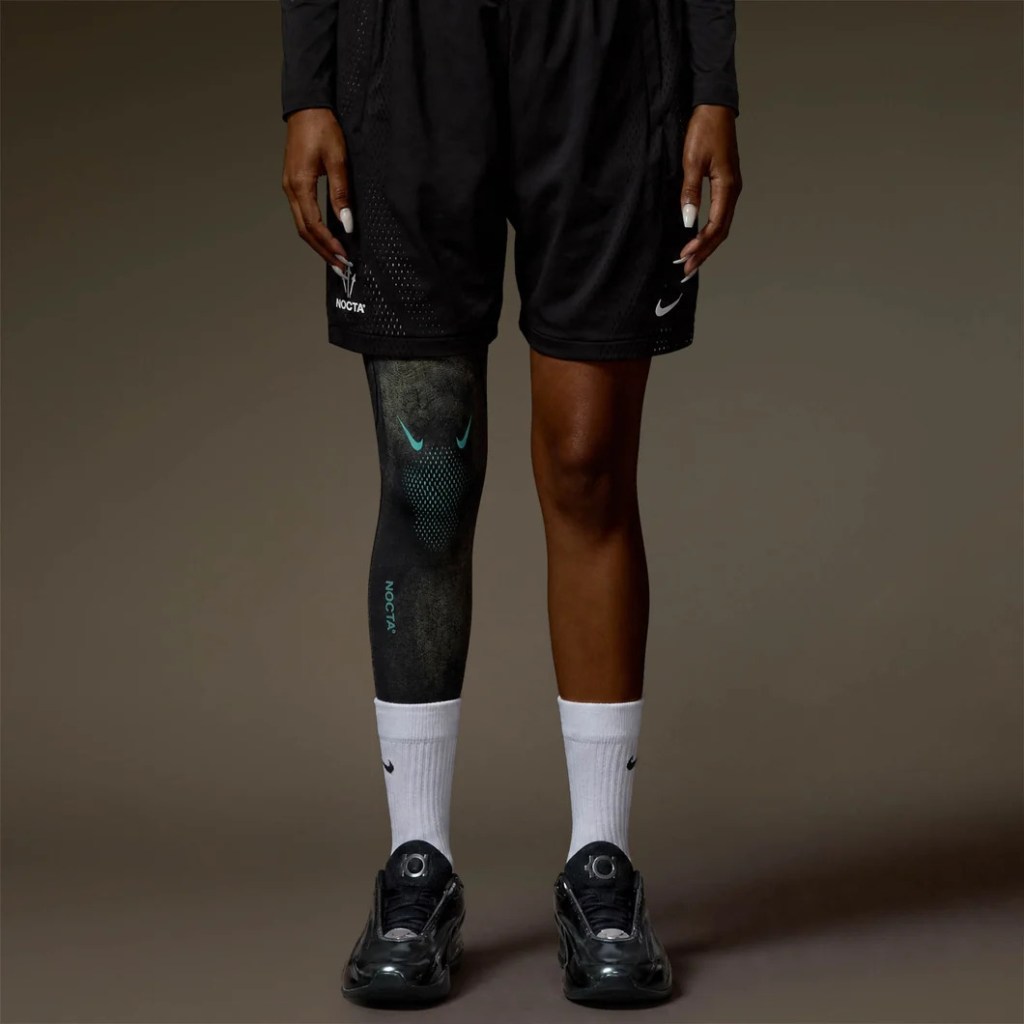 A person wearing black Nike athletic shorts and socks, with a Nike Nocta logo on the leg, standing against a dark background.