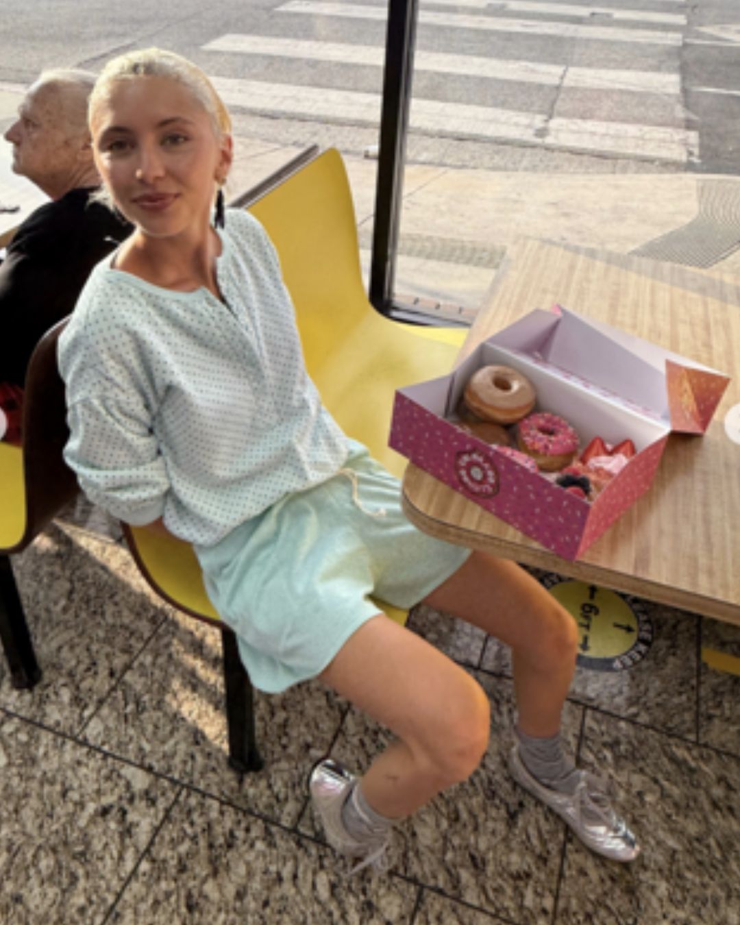 Iris Law sits at a yellow table in front of a box of donuts wearing a blue long-sleeve top with silver trainers and shorts.