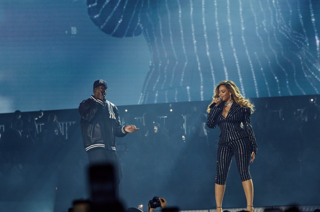 Two performers on stage, a man in a Yankees baseball cap and a woman in a black and white striped jumpsuit, singing and dancing under a large video screen.