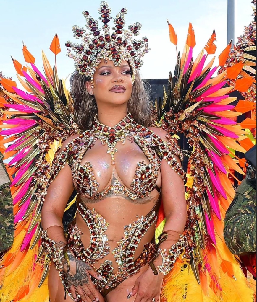 A person wearing an elaborate, colorful costume with feathered headdress and jeweled bra top, posing in a dramatic stance.