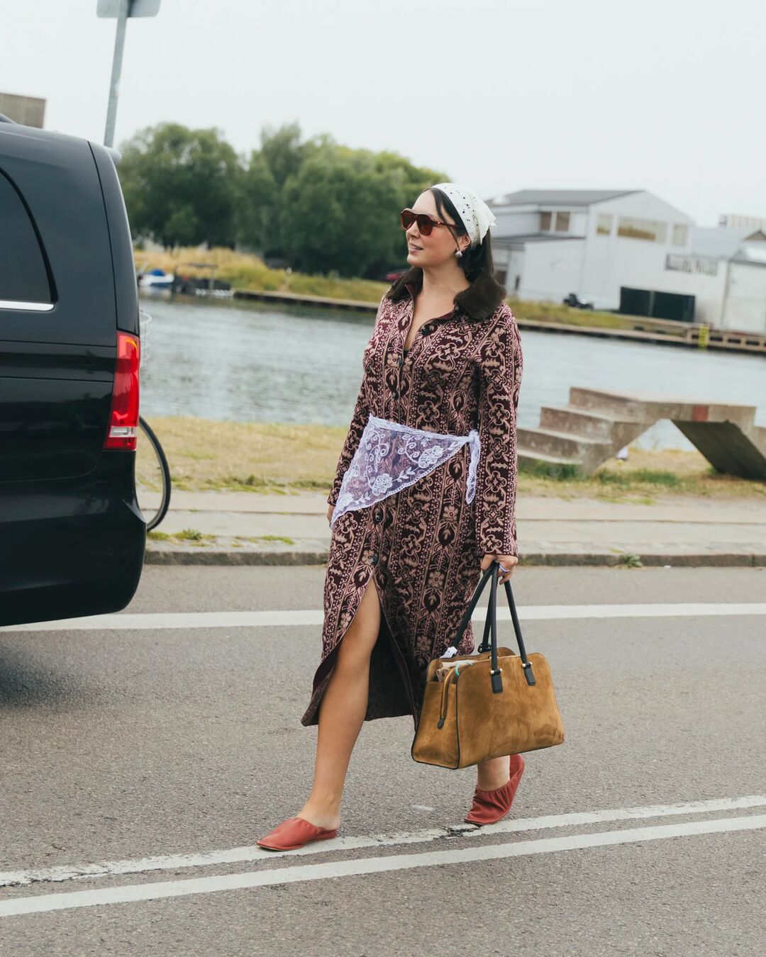 Woman wears dress with scarf around her waist with a suede tote and mules.